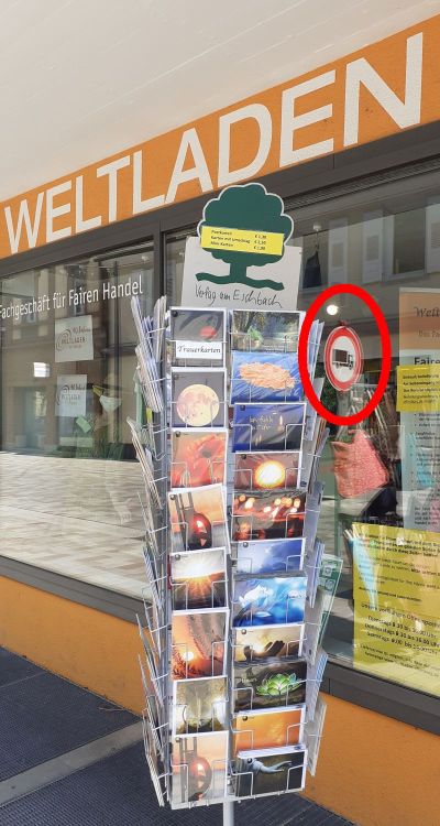Foto: ein Gestell mit Postkarten vor dem Schaufenster des Weltladen Regentropfen. Im Schaufenster spiegelt sich ein Schild, welches die Durchfahrt für Lkws verbietet.
