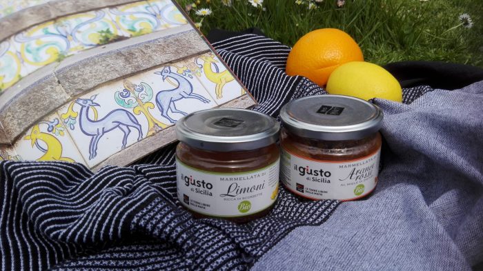 Gläser Zitronen- und Orangenmarmelade von Libera Terra auf Decke mit Orange, Zitrone und Buch mit Abbildung von italienischen Fliesen